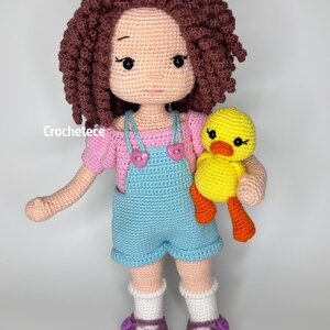 May include: A crocheted doll with brown curly hair, wearing a pink shirt, blue dungarees, and white socks. The doll is holding a yellow crocheted duck with orange feet.