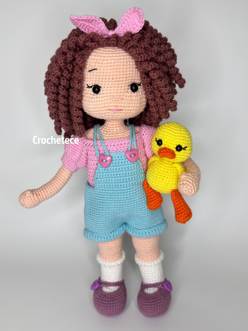 May include: A crocheted doll with brown curly hair, wearing a pink shirt, blue dungarees, and white socks. The doll is holding a yellow crocheted duck with orange feet.