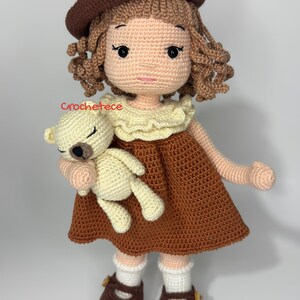 May include: A crocheted doll with brown hair, wearing a brown dress and a brown hat, holding a crocheted teddy bear. The doll is wearing brown shoes with white socks.