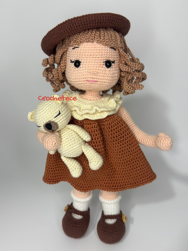 May include: A crocheted doll with brown hair, wearing a brown dress and a brown hat, holding a crocheted teddy bear. The doll is wearing brown shoes with white socks.