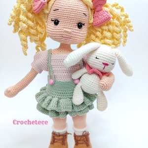 May include: A crocheted doll with yellow curly hair, wearing a pink and green dress, holding a white crocheted bunny. The doll is wearing brown shoes and has pink bows in her hair. The text "Crochetece" is visible in the bottom left corner of the image.