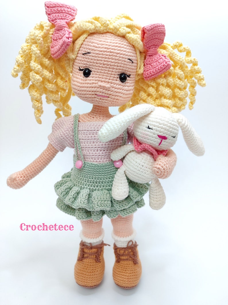May include: A crocheted doll with yellow curly hair, wearing a pink and green dress, holding a white crocheted bunny. The doll is wearing brown shoes and has pink bows in her hair. The text "Crochetece" is visible in the bottom left corner of the image.