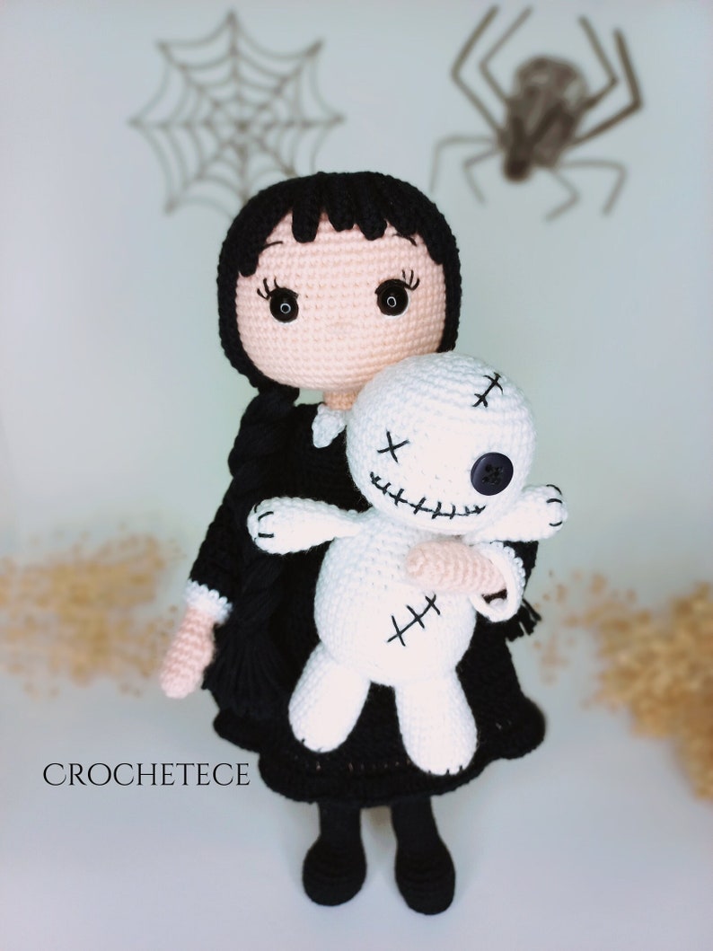 May include: A crocheted doll with black hair and a black dress is holding a white crocheted doll with stitched-on eyes and a stitched-on mouth. The doll is wearing a white collar and black shoes.