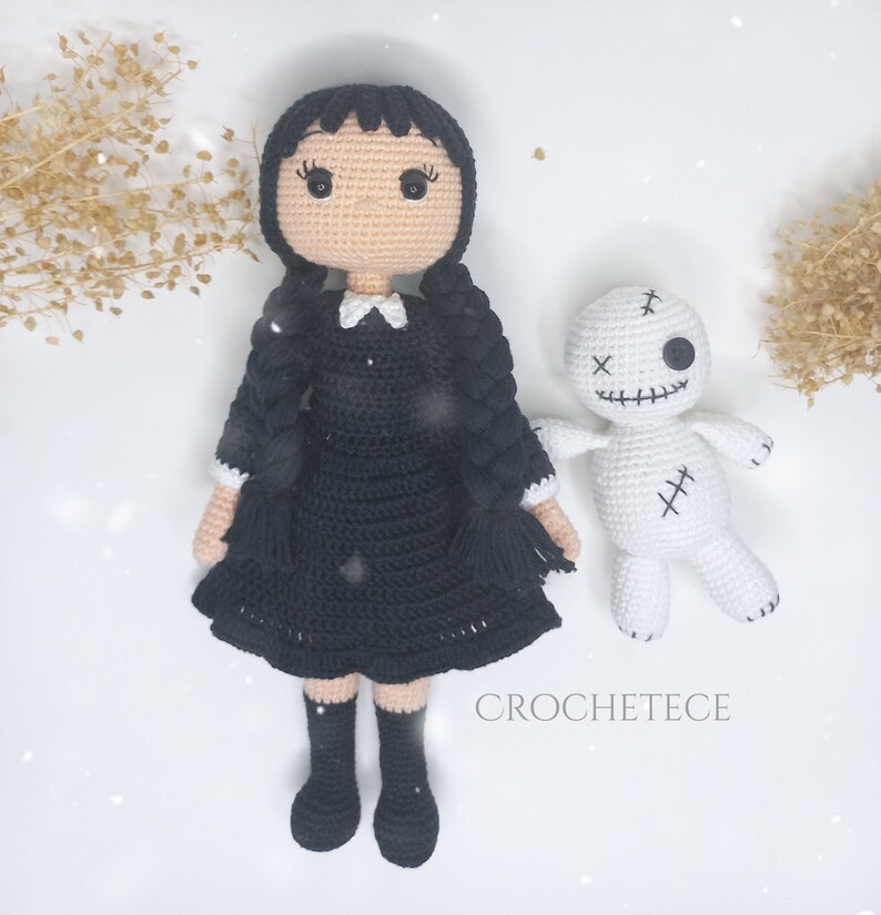 May include: A black and white crocheted doll with black hair in pigtails wearing a black dress and white collar. The doll is standing next to a white crocheted voodoo doll with black stitching and a single black button eye.
