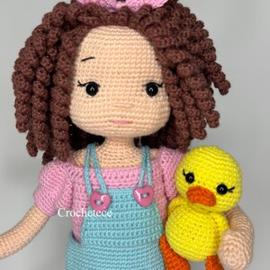 May include: A crocheted doll with brown curly hair, wearing a pink shirt and blue dungarees, holding a yellow crocheted duck. The doll has a pink bow in her hair and pink buttons on her dungarees. The text "Crochetece" is visible on the doll's dungarees.