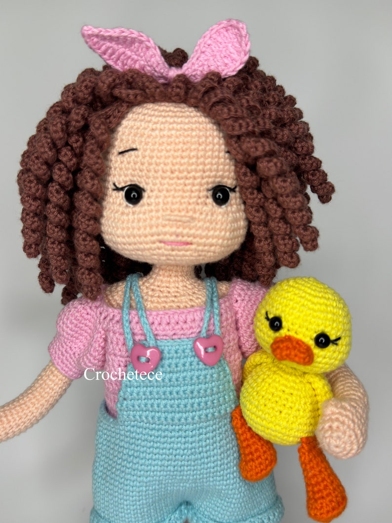 May include: A crocheted doll with brown curly hair, wearing a pink shirt and blue dungarees, holding a yellow crocheted duck. The doll has a pink bow in her hair and pink buttons on her dungarees. The text "Crochetece" is visible on the doll's dungarees.