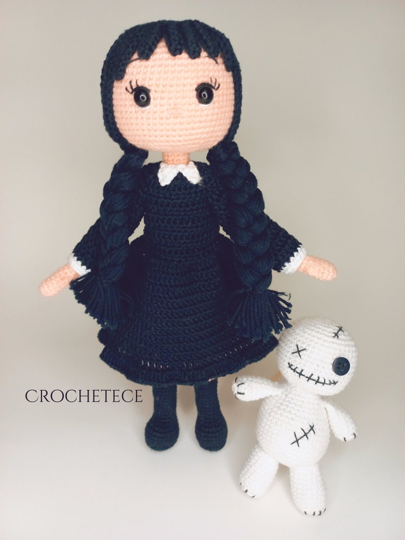 May include: A crocheted doll with black hair in pigtails wearing a black dress and white collar, holding a white crocheted doll with black stitching and a button eye. The dolls are on a white background. CROCHETETECE is written in the bottom left corner.