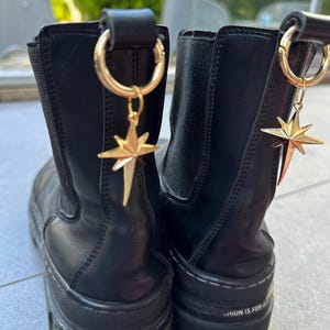 May include: Pair of black leather ankle boots with gold star charms attached to the top. The boots have a chunky platform sole and the text "Fashion is for brave people" is printed on the sole of one boot.