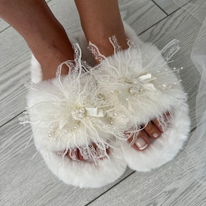 May include: White fluffy slippers with a delicate lace and pearl embellishment. The slippers have a bow and a fluffy pom-pom.