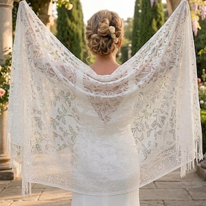 Puede incluir: Un chal de encaje blanco con un diseño floral se muestra, revelando un vestido blanco. El chal tiene flecos en los bordes. El fondo incluye un arco con flores y vegetación, lo que sugiere una boda o un evento formal.