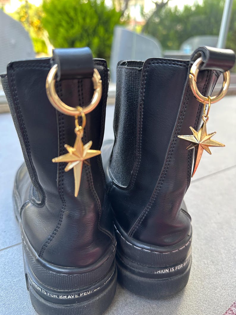 May include: Pair of black leather ankle boots with gold star charms attached to the top. The boots have a thick platform sole and the text "Fashion is for brave people" is printed on the side of each boot.