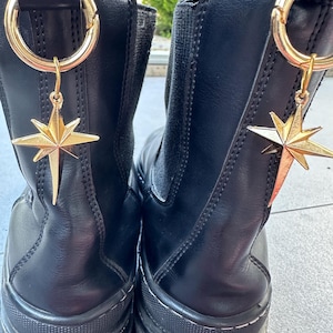 May include: A pair of black leather ankle boots with gold star charms attached to the top of each boot. The boots have a thick rubber sole and the text "Fashion is for brave people" is printed on the bottom of the boots.
