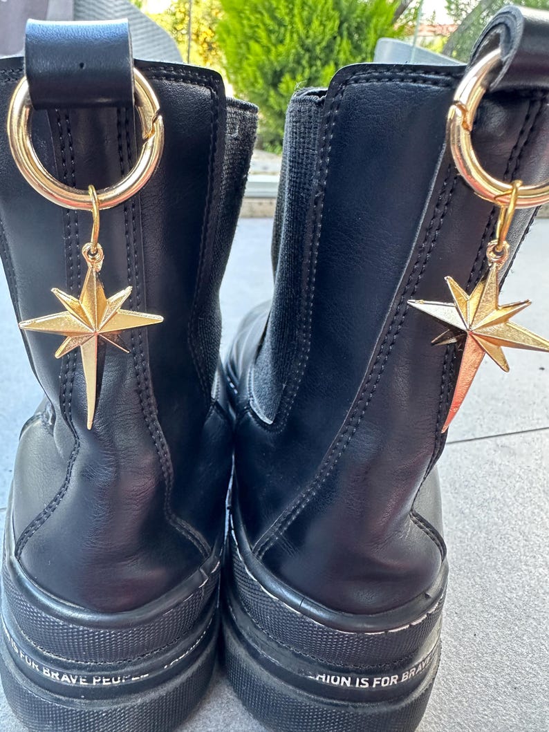 May include: A pair of black leather ankle boots with gold star charms attached to the top of each boot. The boots have a thick rubber sole and the text "Fashion is for brave people" is printed on the bottom of the boots.