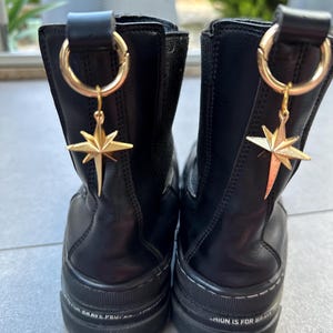 Puede incluir: Un par de botines negros de cuero con suela de plataforma gruesa. Cada bota tiene un dije de estrella dorada unido a un anillo en la parte posterior de la bota. El texto "Fashion is for brave souls" está impreso en la suela de una bota, y el texto "Never give up" está impreso en la suela de la otra bota.