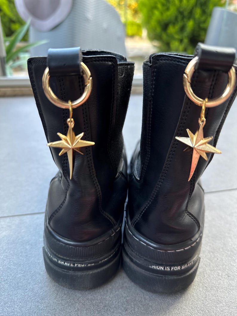 May include: A pair of black leather ankle boots with a chunky platform sole. Each boot has a gold star charm attached to a ring on the back of the boot. The text "Fashion is for brave souls" is printed on the sole of one boot, and the text "Never give up" is printed on the sole of the other boot.