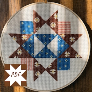 Puede incluir: Un bastidor de bordado de punto de cruz con un diseño de estrella. El diseño incorpora formas geométricas marrones, azules y rosas con pequeños detalles de flores blancas. El texto "PDF" está en una forma de estrella marrón en la esquina inferior izquierda. El bastidor está sobre un fondo de madera.
