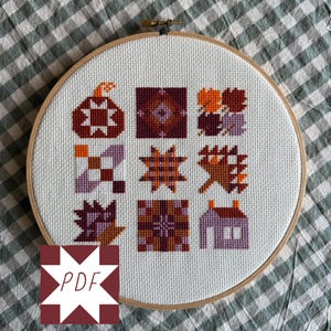 Könnte beinhalten: Ein runder Holzstickrahmen mit weißem Aida-Stoff, der ein Kreuzstichmuster mit neun verschiedenen Quadraten zeigt. Die Motive umfassen einen Kürbis, geometrische Quiltblöcke, Herbstblätter und ein kleines Haus in den Farben Orange, Lila und Kastanienbraun.