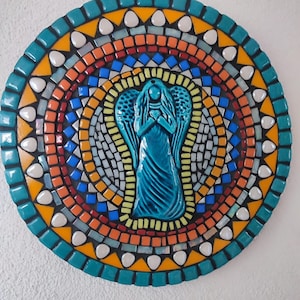 Puede incluir: Un arte de pared redondo de mosaico con una figurita de ángel turquesa en el centro. El mosaico está hecho de pequeñas baldosas de colores en tonos de azul, verde, amarillo, naranja y rojo. El ángel tiene alas y tiene las manos juntas en oración.