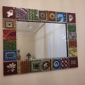 Könnte beinhalten: Ein quadratischer Spiegel mit einem Mosaikrahmen aus bunten Fliesen. Der Rahmen zeigt verschiedene Designs, darunter eine Taube, eine Blume, einen Elefanten und ein böses Auge. Der Spiegel ist von einem dunkelbraunen Rand umgeben.