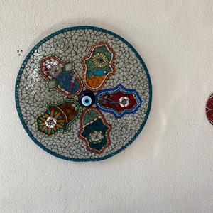 Puede incluir: Un arte de pared redondo de mosaico con un fondo blanco y azulejos de mosaico coloridos. El arte presenta cinco manos de hamsa, cada una con un diseño diferente. El centro del arte presenta un amuleto de ojo azul.