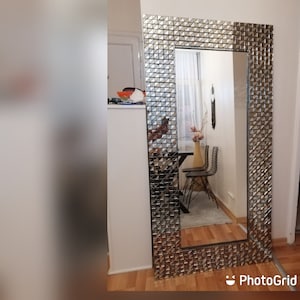 Puede incluir: Un espejo grande y rectangular con un marco plateado hecho de pequeñas baldosas geométricas. El espejo refleja una habitación con una ventana, una silla y una mesa.