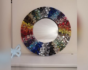 May include: A round mirror with a colorful mosaic frame. The frame is made of small, square tiles in shades of red, orange, yellow, green, blue, and purple. The mirror is surrounded by a black border.