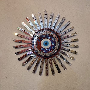 Puede incluir: Un adorno de pared decorativo con un diseño de mosaico que presenta un ojo azul maligno en el centro. El mosaico está rodeado por un anillo de azulejos de espejo plateados.