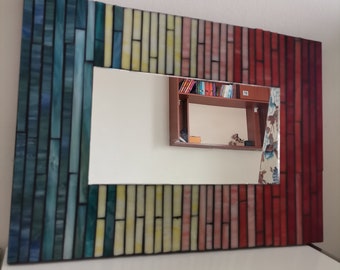 May include: A rectangular mirror with a colorful mosaic frame. The frame is made of small, rectangular tiles in shades of blue, green, yellow, pink, and red. The mirror is reflecting a bookshelf with books.