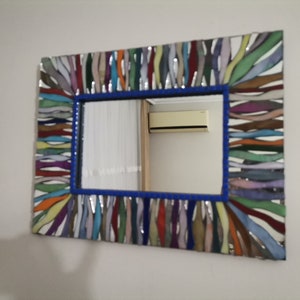 Könnte beinhalten: Ein rechteckiger Spiegel mit einem bunten Mosaikrahmen. Der Rahmen besteht aus kleinen, bunten Glasfliesen, die in einem wellenförmigen Muster angeordnet sind. Der Spiegel ist von einem blauen Rand umgeben.