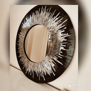 May include: A circular mirror featuring a black mosaic tile surround and silver, linear embellishments extending outwards. The mirror's reflective surface is visible within the frame. The design is contemporary and artistic, suitable for various interior styles.