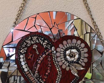 May include: A round mirror with a mosaic design featuring a red heart with silver and white accents. The mirror is hanging from a silver chain.