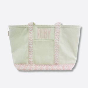 May include: A light green canvas tote bag with pink accents. The word "RUBY" is embroidered in pink on the front pocket. The handles and bottom trim feature a pink and white leaf pattern. A small tag is visible on the side.