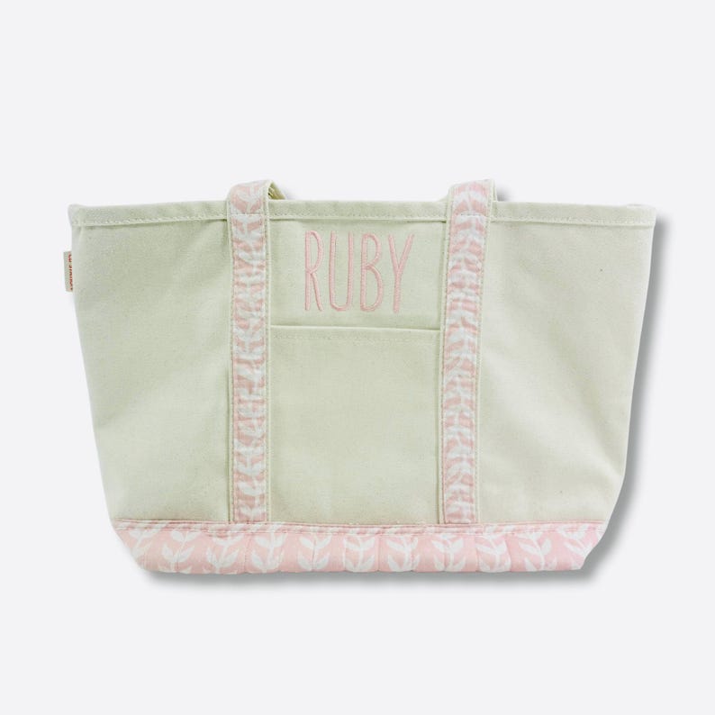 May include: A light green canvas tote bag with pink accents. The word "RUBY" is embroidered in pink on the front pocket. The handles and bottom trim feature a pink and white leaf pattern. A small tag is visible on the side.
