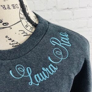 May include: A dark gray sweatshirt with a light blue embroidered design that says "Laura Co. Rae".