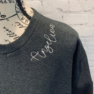 May include: A dark gray sweatshirt with the name "Angelica" embroidered in white on the shoulder.