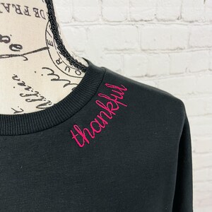 May include: Black sweatshirt with a pink embroidered word "thankful" on the neckline.