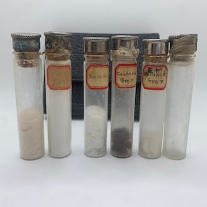 Puede incluir: Un juego de seis pequeños frascos de vidrio con tapas plateadas y etiquetas. Las etiquetas son blancas con letras rojas e incluyen las palabras "Ácido bórico", "Cascara Grs." y "Aspirina Grs."