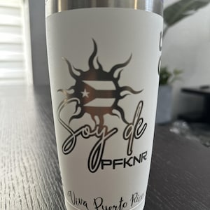 May include: White insulated tumbler with a silver lid. The tumbler features a black graphic of the Puerto Rican flag within a sunburst design, with the words "Soy de PFKNR" and "Viva Puerto Rico" in black script.