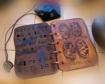 May include: A copper-colored journal with a ring binding. The front cover has a variety of symbols and the back cover has four oval-shaped images of people. The journal is open to reveal the inside pages, which are also covered in symbols.