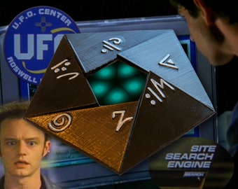 May include: A black, six-sided, pyramid-shaped object with a glowing green light inside. The object has symbols on each side, including a circle with a dot in the center, a number 7, a symbol that looks like a backwards 'M', a triangle, a symbol that looks like a backwards 'P', and a symbol that looks like a backwards '2'.