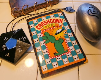 May include: A black and blue dice with symbols on each side, a silver alien spaceship, and a notebook with the words "Crashdown Cafe" and a cartoon cactus on the cover.
