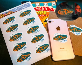 May include: A sheet of stickers with the logo "Crash Down Cafe" in a retro style. The logo features a stylized font with a blue and orange color scheme. The stickers are arranged in a grid pattern on a white background.