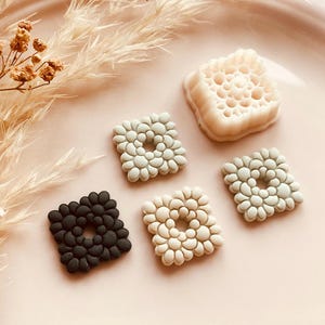 May include: A collection of square-shaped decorative elements in various colors, including black, cream, and light gray, with a textured, pebble-like design. The elements are arranged on a pink surface, with dried floral accents in the background.