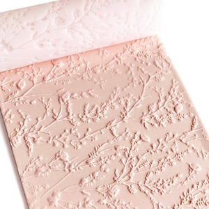 Peut inclure: Un rouleau à pâtisserie rose avec un motif floral en relief. Le design présente des éléments botaniques détaillés, notamment des feuilles et des fleurs, créant une surface texturée. Le rouleau est partiellement déroulé, révélant le motif.