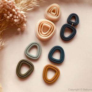 Op de afbeelding: Een verzameling geometrische kleisnijders in verschillende kleuren, waaronder beige, olijfgroen en marineblauw. De snijders zijn gerangschikt op een lichtroze oppervlak, met gedroogde bloemen op de achtergrond. De vormen omvatten geneste driehoeken en verbonden cirkels.