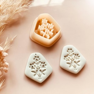 Puede incluir: Un cortador de galletas con forma de flor de color beige claro y dos colgantes de arcilla blanca con diseños florales. El cortador de galletas tiene forma de ramo de flores. Los colgantes tienen pequeños detalles plateados. Flores secas en el fondo. Copyright Babycutters 2025.