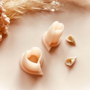 May include: Two heart-shaped cookie cutters and two petal-shaped cutters in a soft, off-white colour. The cutters are arranged on a light pink surface, with dried floral elements in the background. Copyright Babycutters 2025.