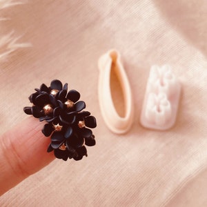 Puede incluir: Un grupo de cuentas en forma de flor azul oscuro con detalles dorados. También se ven dos cortadores de galletas beige. La imagen está sobre un fondo suave de color melocotón. Copyright Babycutters, All Rights Reserved 2023.