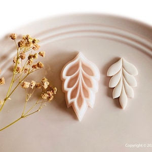 May include: Two leaf-shaped cookie cutters. One is a larger, layered design with a light pink interior, and the other is a smaller, solid white leaf shape. Both are on a light pink plate with dried flowers.