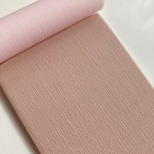 May include: A roll of pale pink crepe paper, partially unrolled to show its textured surface. The paper has a subtle, vertical grain, and the colour is a soft, muted pink. The background is a plain white surface.